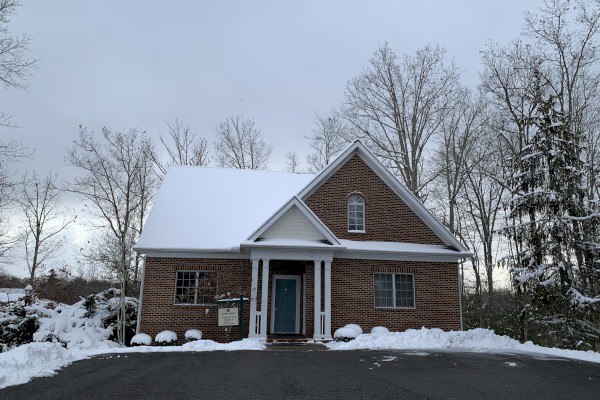 A brick house with a snow-covered roof and driveway, bare trees in the background, and a light dusting of snow around the yard.