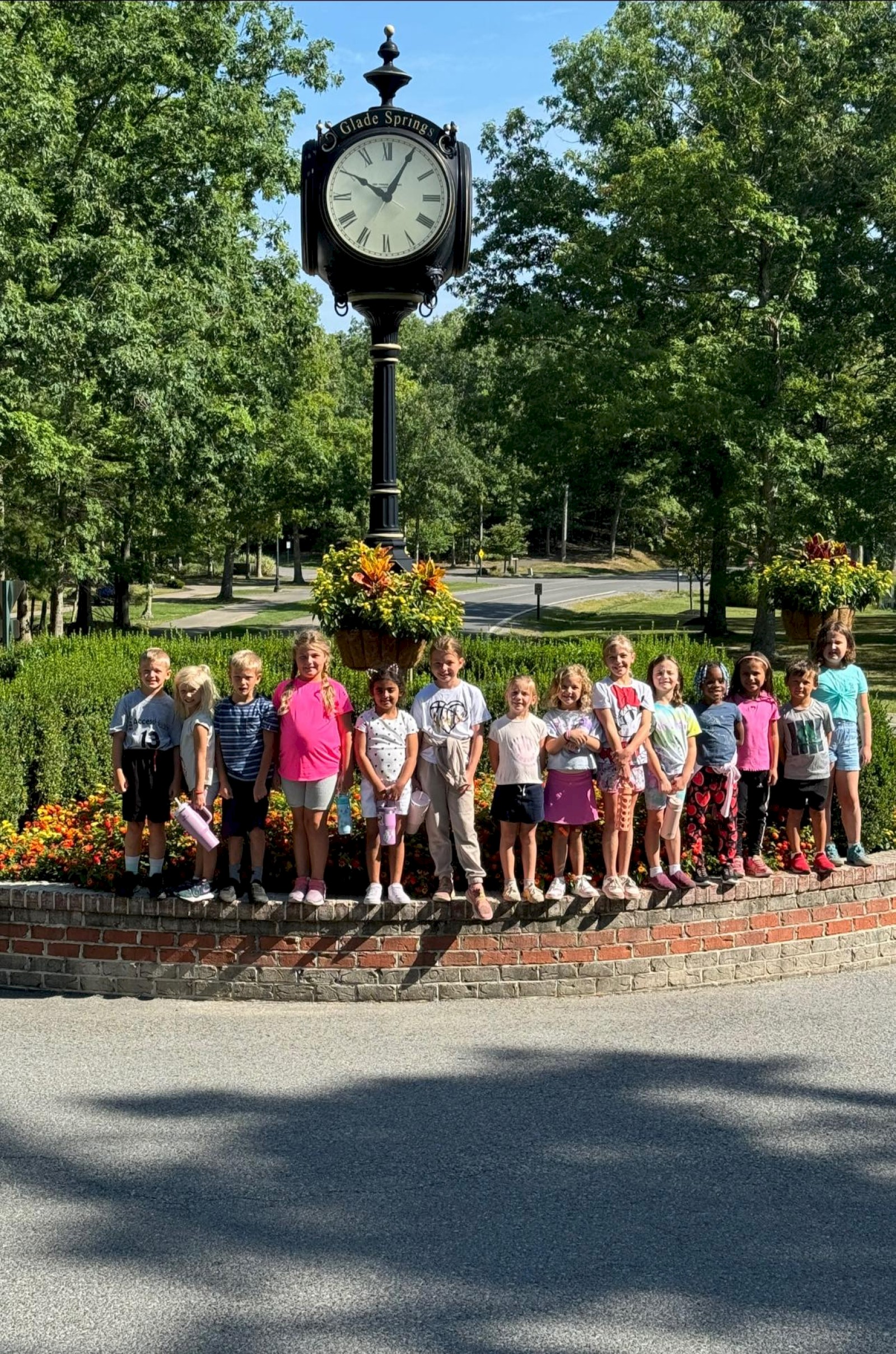 A group of children stands in a line around a brick planter with a tall clock post, trees and flowers behind, sunny day.