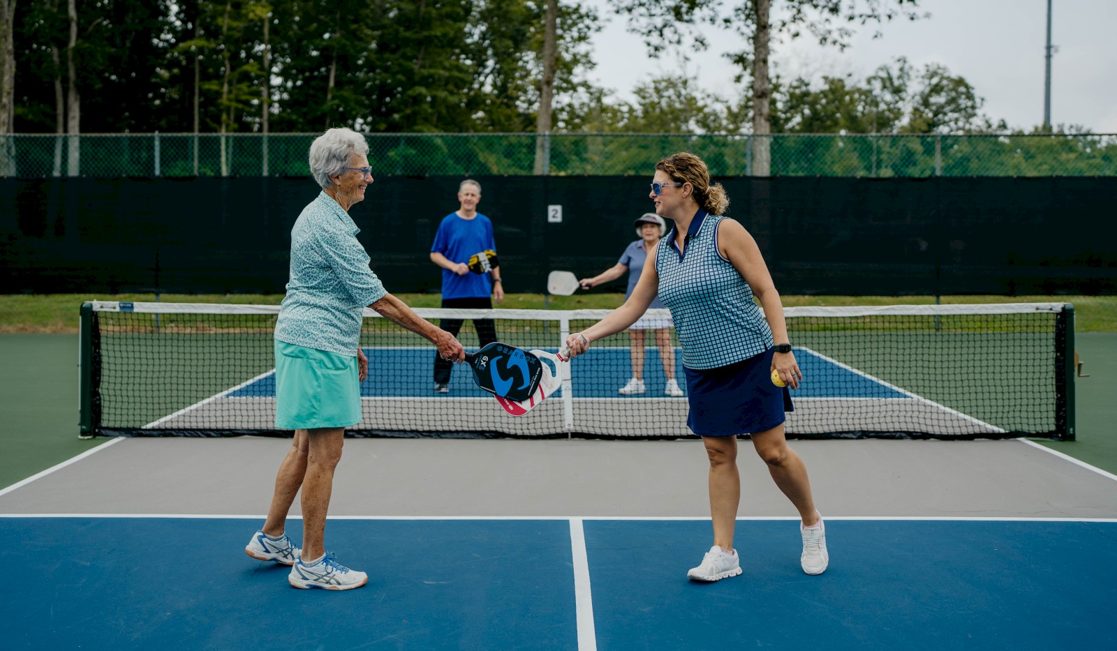Two people on a pickleball court are exchanging a paddle at the net, with two others in the background.