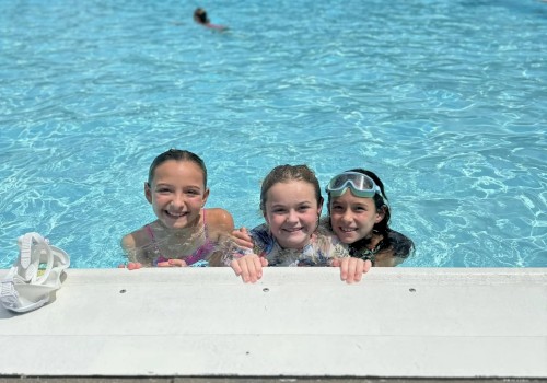 Three kids grin from the pool edge as the sunny day shines, with lounge chairs and umbrellas in the background, enjoying a splashy moment.