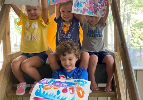 Three kids on a wooden playset hold up colorful paintings; a younger child sits in front with a bright blue artwork, smiling joyfully.