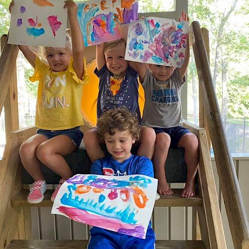 Three kids on a wooden playset hold up colorful paintings; a younger child sits in front with a bright blue artwork, smiling joyfully.