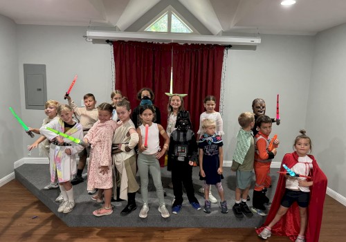 A group of children in a stage-like room wearing costumes, some holding glow sticks or toy swords, posing for a photo with a red curtain backdrop.