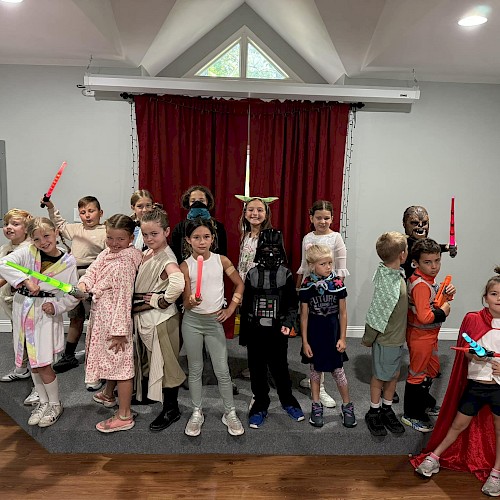 A group of children in a stage-like room wearing costumes, some holding glow sticks or toy swords, posing for a photo with a red curtain backdrop.