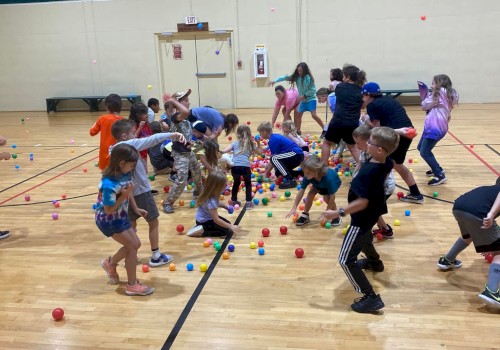 Kids in a gym playing a game with colorful cups scattered on the floor; they’re laughing and moving around excitedly, indoors.