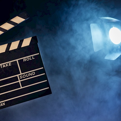 A behind-the-scenes film set scene: a clapperboard in hand, studio lights, and smoky atmospheric glow ready for filming. Ending with a period.