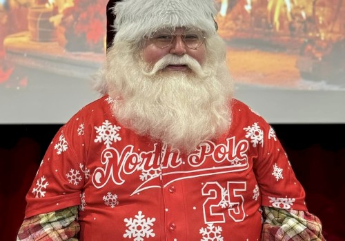 A bearded man in a red Christmas sweater sits in front of a fireplace backdrop, wearing a Santa hat and festive 25th North Pole gear.