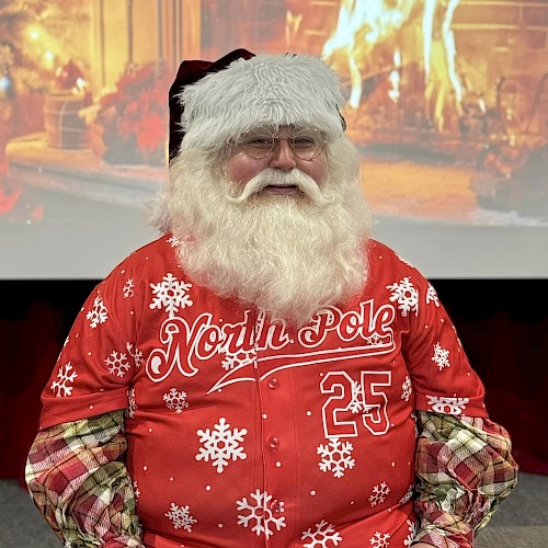 A bearded man in a red Christmas sweater sits in front of a fireplace backdrop, wearing a Santa hat and festive 25th North Pole gear.