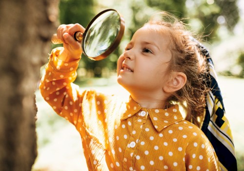 A young girl in a polka-dot orange shirt uses a magnifying glass outdoors, exploring energy and curiosity in a sunny park.