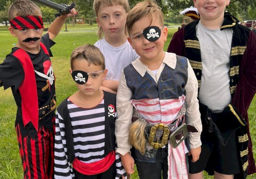 A group of five kids dressed in pirate costumes posing on a grassy field, some with eye patches and swords, smiling at the camera.