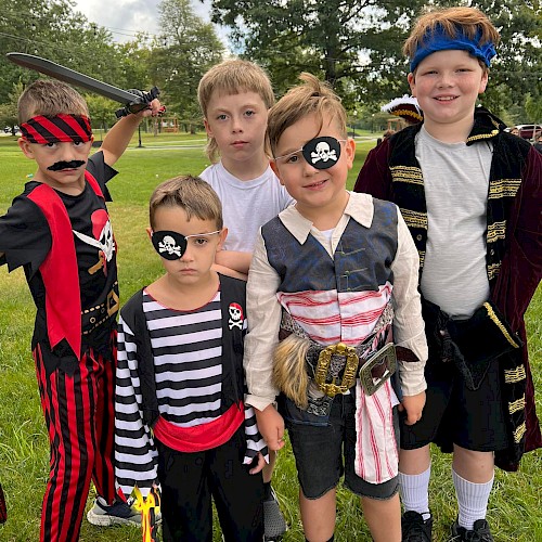 A group of five kids dressed in pirate costumes posing on a grassy field, some with eye patches and swords, smiling at the camera.