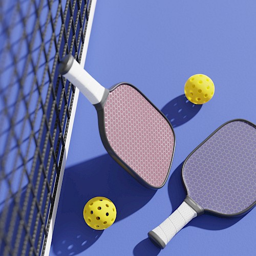 Two pickleball paddles and three yellow balls are on a blue court next to a net.
