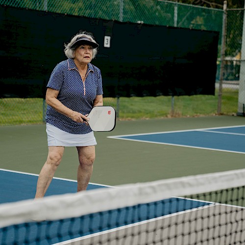 A person is playing pickleball, wearing a visor and holding a paddle on an outdoor court near a net, focusing on the game.