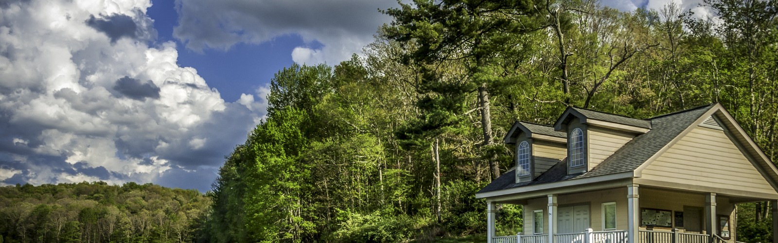 A serene lakeside cabin sits by calm water, lush trees, and a dramatic sky filled with fluffy clouds.