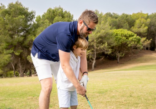 An adult is teaching a child how to play golf on a grassy course, with trees in the background, under a clear sky.
