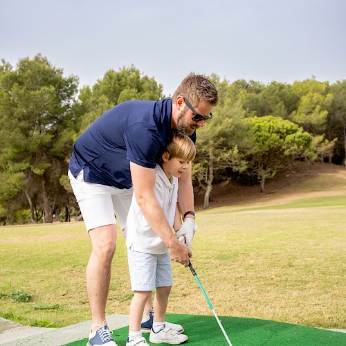 An adult is teaching a child how to play golf on a grassy course, with trees in the background, under a clear sky.