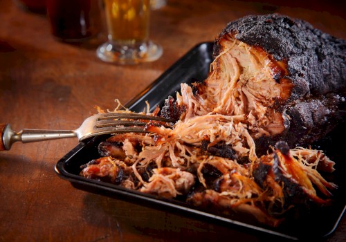 A serving of shredded pulled pork with a barbecue crust sits in a black tray, accompanied by a fork on a wooden table.
