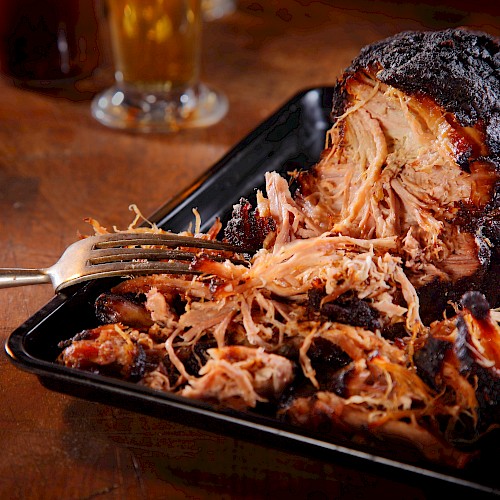 A serving of shredded pulled pork with a barbecue crust sits in a black tray, accompanied by a fork on a wooden table.