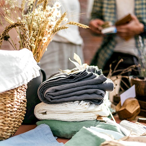 A display featuring folded linens, a basket with dried flowers, and a person in the background holding an item, outdoors.