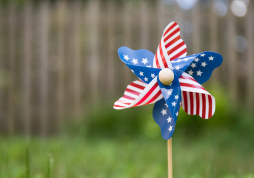 A pinwheel with a red, white, and blue star pattern stands in the grass, with a wooden fence blurred in the background.
