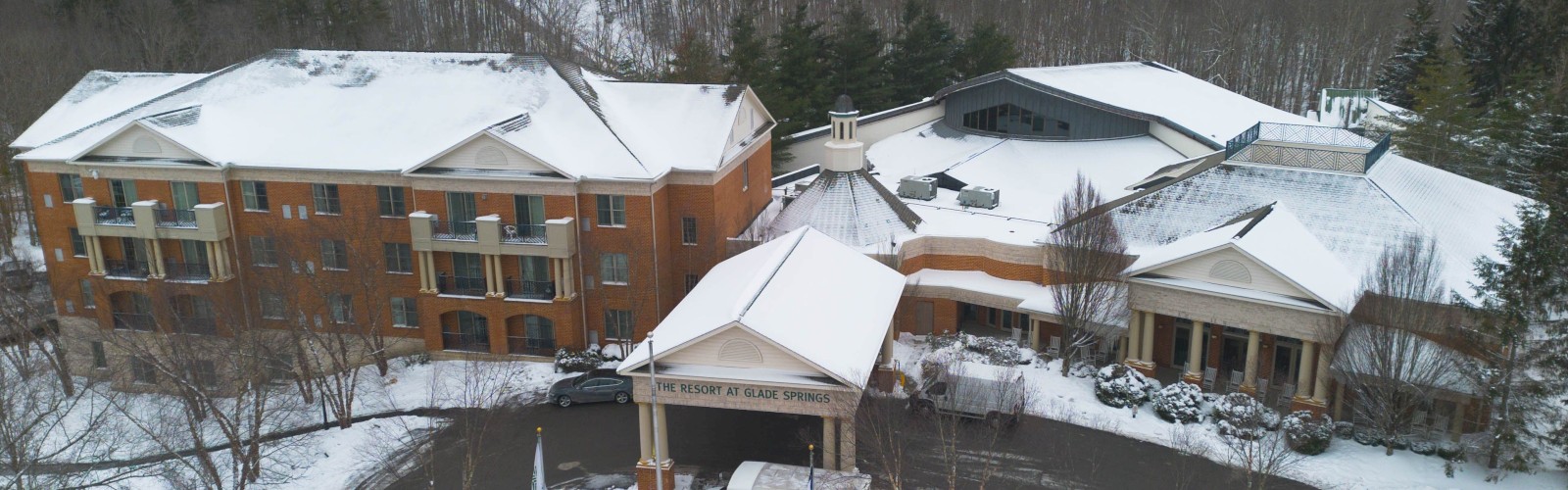 Snowy resort or condo complex with brick buildings, a central covered entrance, a circle driveway, and snow-covered trees surrounding it.