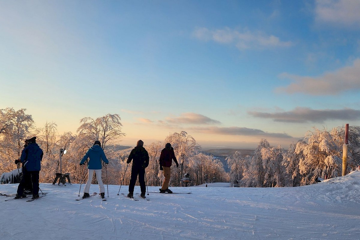 Skier group on a snowy slope at sunset, trees frosted white, chairlift on the right, calm blue sky, tranquil winter scene.