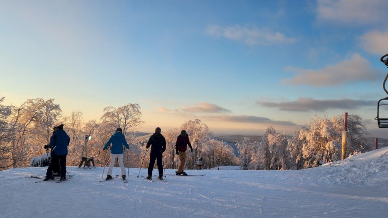 Skier group on a snowy slope at sunset, trees frosted white, chairlift on the right, calm blue sky, tranquil winter scene.