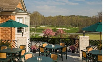 An outdoor patio with tables, chairs, umbrellas, flowers, and a scenic view of a golf course and trees in the background.