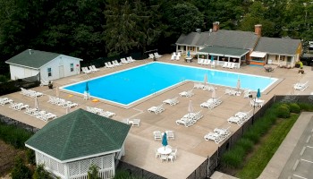 An outdoor swimming pool surrounded by white lounge chairs, umbrellas, and a clubhouse in a green, wooded area.