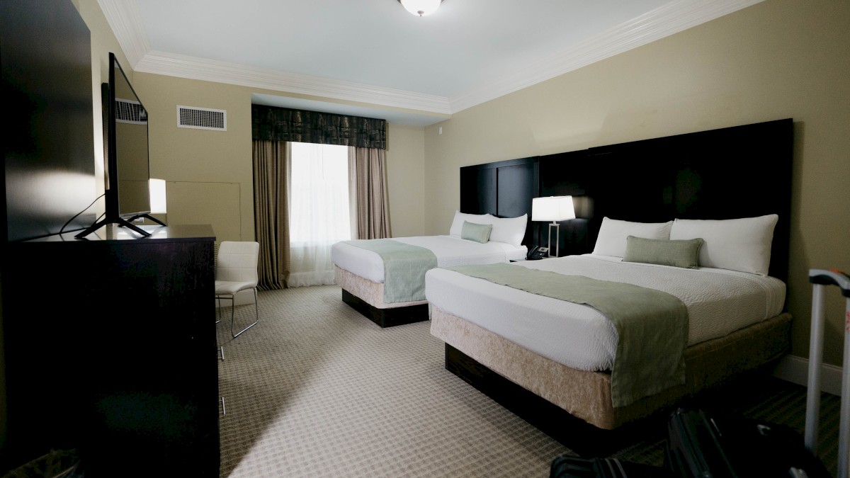 A hotel room with two beds, a TV, nightstand, lamps, chairs, carpet, and curtains in neutral tones.