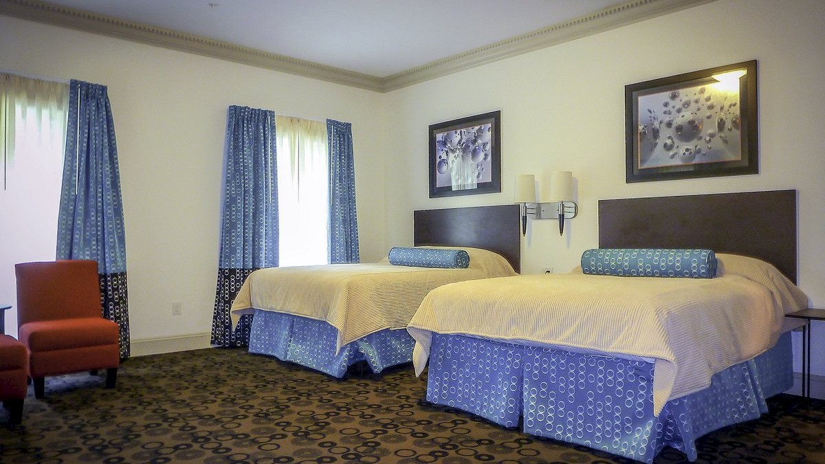 A hotel room with two beds, blue curtains, framed pictures, a red chair, and patterned carpet, all under a neutral wall background.