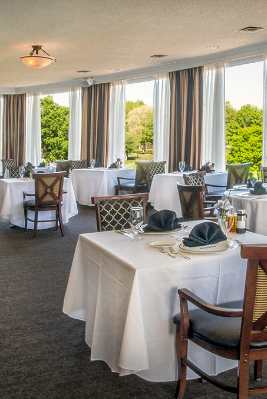 This is a brightly lit dining room with elegant tables set with white tablecloths, napkins, glasses, and silverware, overlooking a lush green landscape.