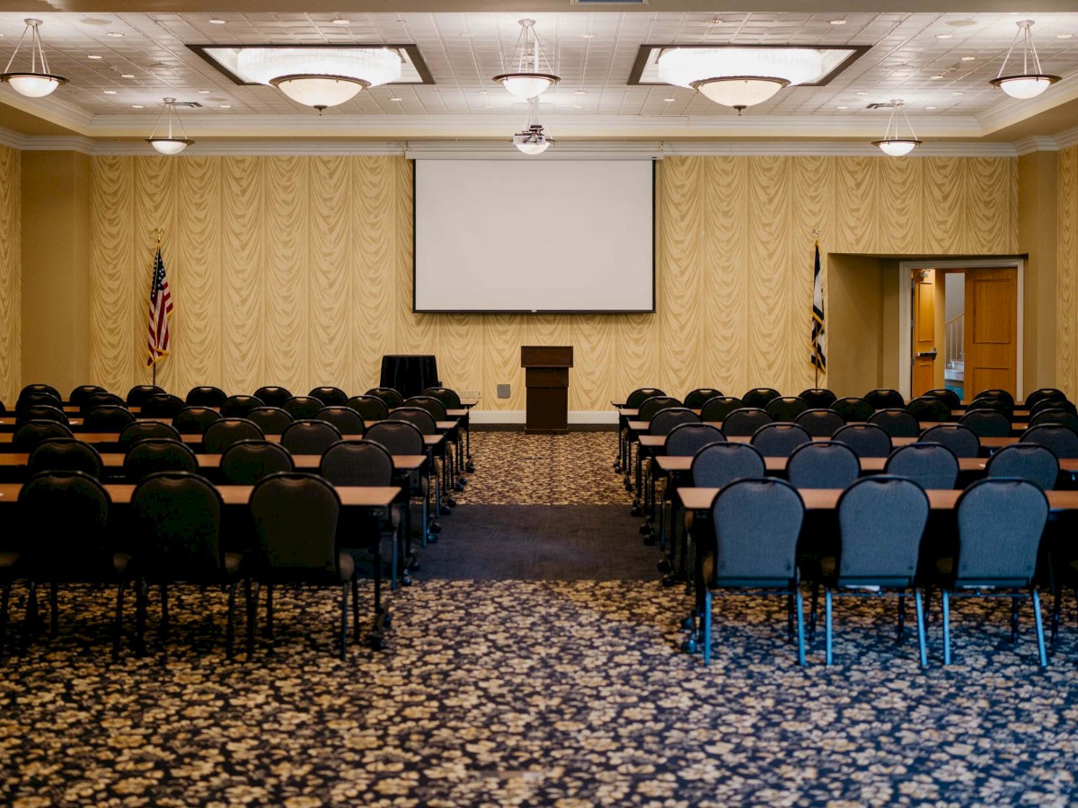 A conference room with chairs, tables, a large screen, flags, and a podium, set up for a presentation or meeting.