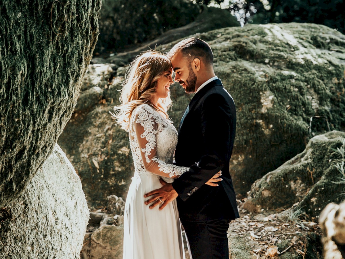 A couple in wedding attire share an intimate moment outdoors, standing between large mossy rocks in a natural setting.