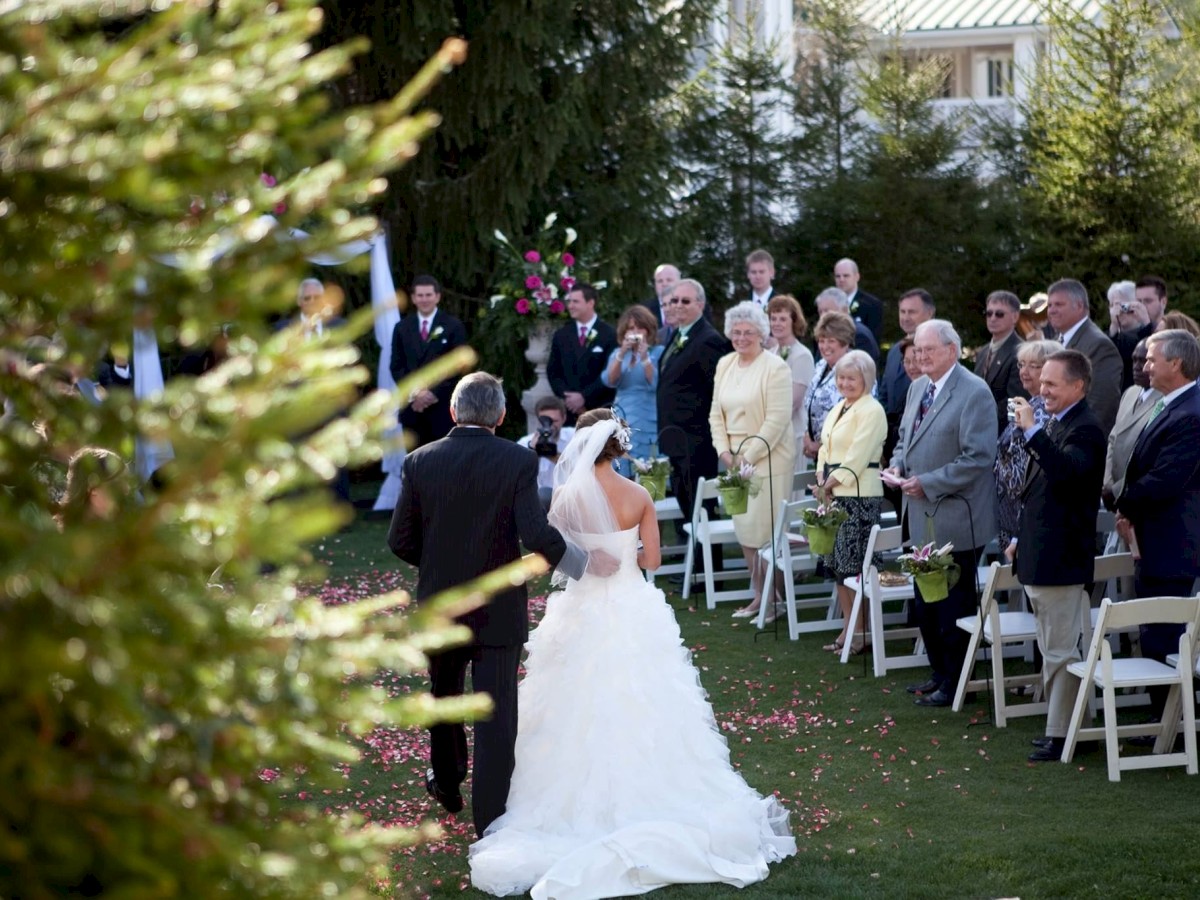 A bride and groom walk down the aisle at an outdoor wedding ceremony with guests standing and smiling, surrounded by greenery and trees.