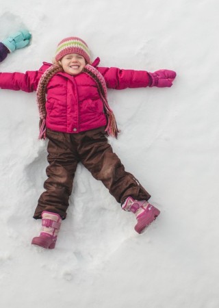 Two young girls are lying in the snow, smiling and making snow angels while dressed in colorful winter clothing.