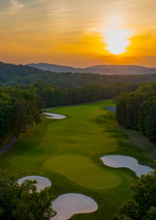 A scenic golf course at sunset surrounded by lush trees and rolling hills, with sand bunkers and a distant mountain range.