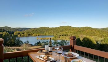 A scenic view with a lake and hills, featuring a wooden table set with gourmet food, wine, and glasses, outdoors under a clear blue sky.
