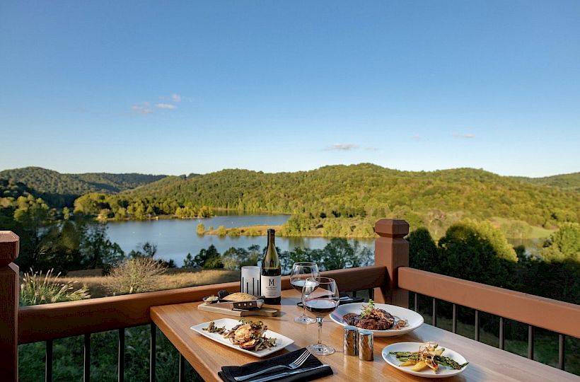 A scenic view with a lake and hills, featuring a wooden table set with gourmet food, wine, and glasses, outdoors under a clear blue sky.