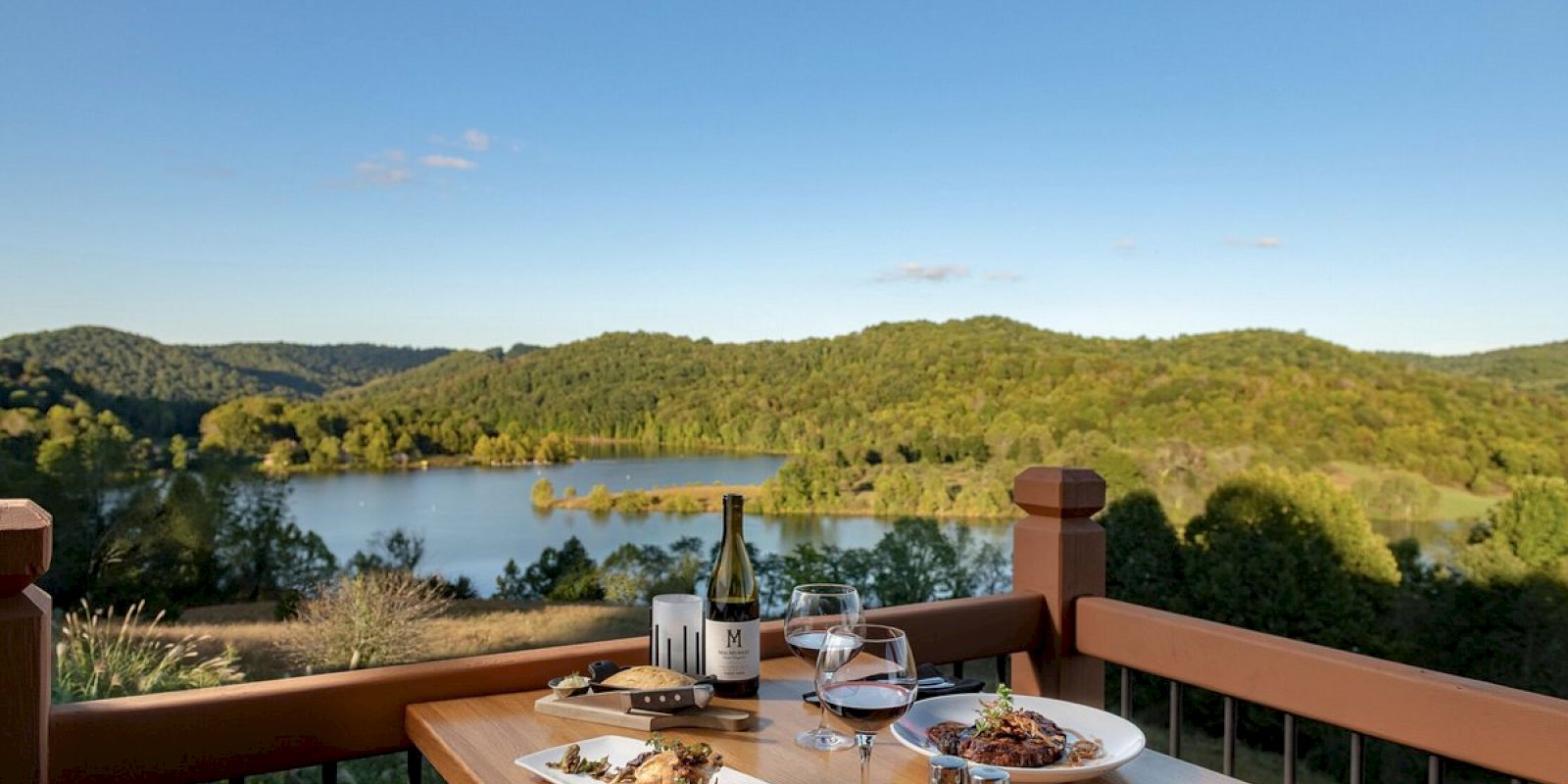 A scenic outdoor dining setup with two plates of food, a bottle of wine, and glasses overlooking a lake and lush green hills.