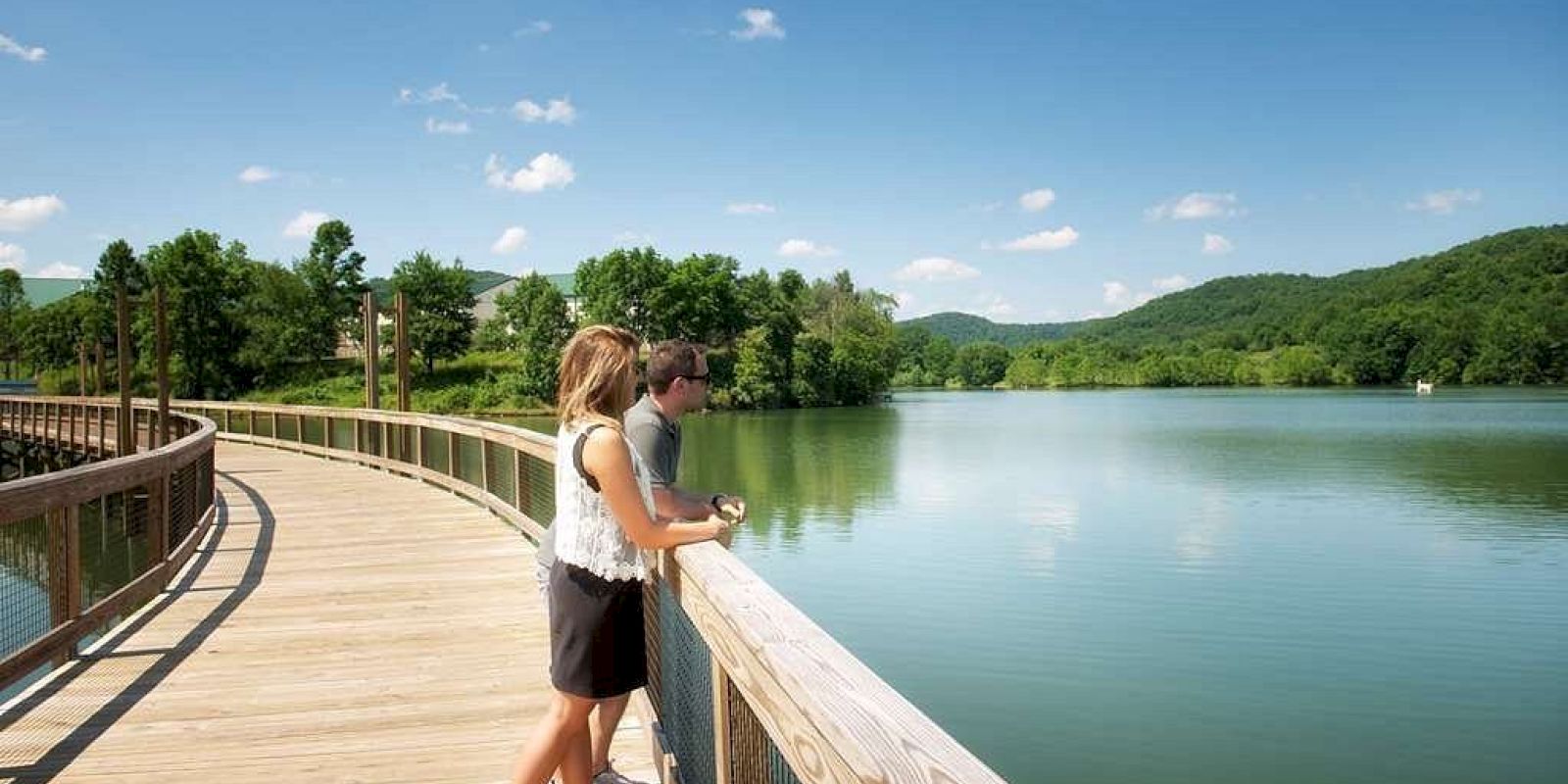 A couple is standing on a wooden walkway overlooking a serene lake surrounded by green hills and a clear blue sky.
