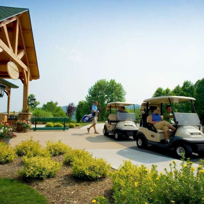 People are driving golf carts near a building with landscaped surroundings and trees in the background, on a sunny day.