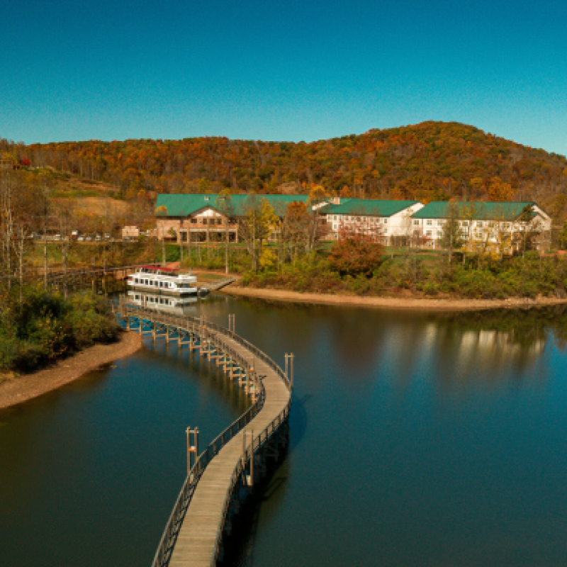 A serene lake scene with a winding boardwalk, a docked boat, and buildings surrounded by autumn trees and hills under a clear blue sky.