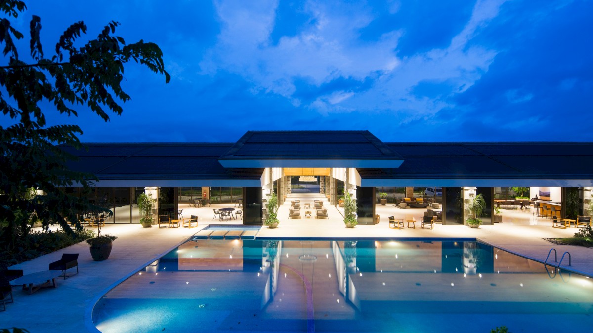 A luxurious modern building with a large illuminated swimming pool in the foreground, surrounded by greenery under a twilight sky.