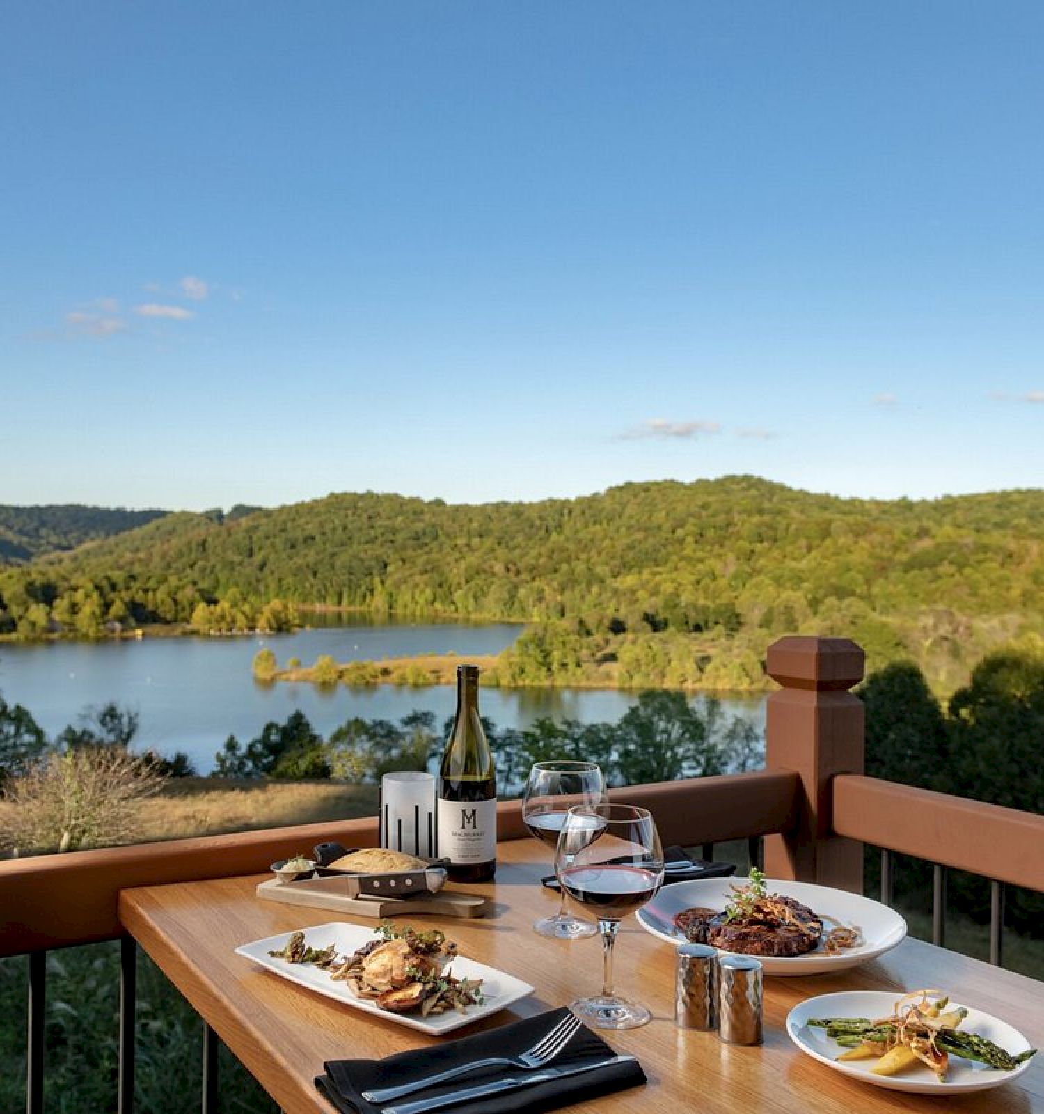 A table set with plates of food and wine overlooks a scenic lake and forested hills under a clear blue sky.