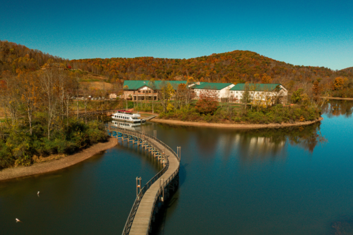A scenic view of a lake with a curved bridge leading to a building complex surrounded by colorful autumn trees and hills in the background.
