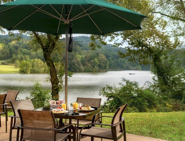 Outdoor dining setup with a table, chairs, and a large green umbrella, overlooking a scenic lake and wooded area.