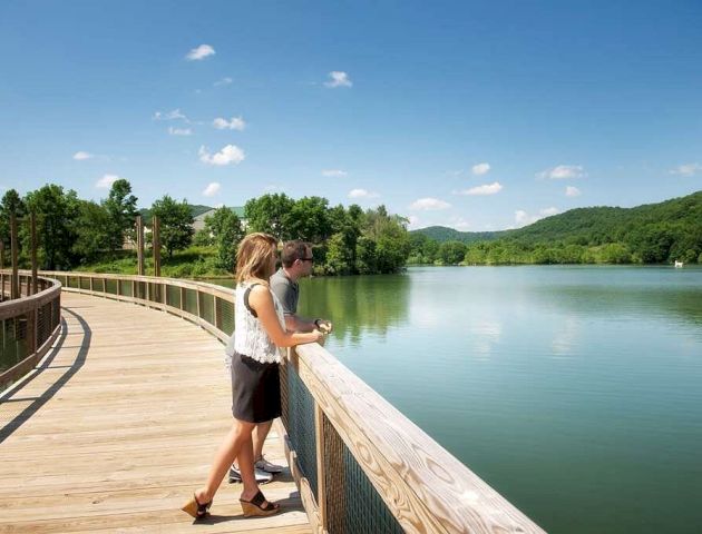 A couple stands on a wooden boardwalk overlooking a serene lake with lush green hills in the background under a clear blue sky.