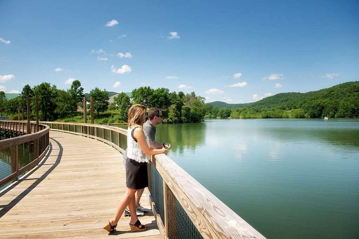 A couple stands on a wooden boardwalk overlooking a lake, surrounded by lush green hills under a clear blue sky.