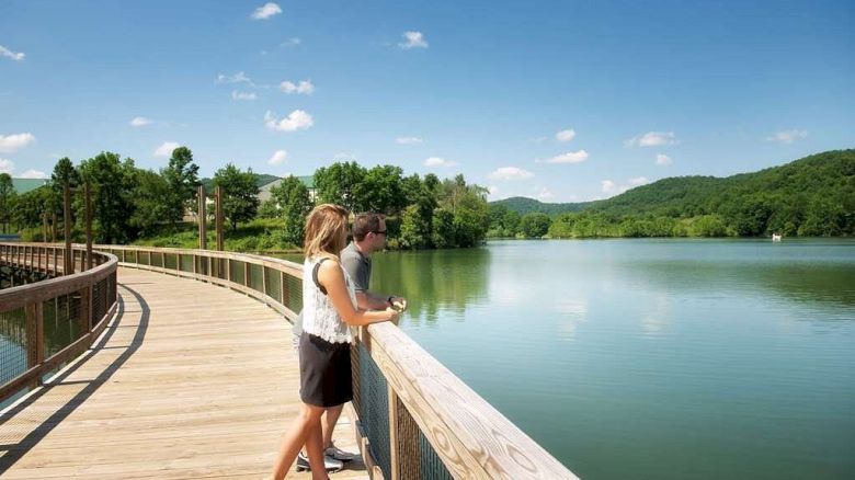A couple stands on a wooden boardwalk overlooking a lake, surrounded by lush green hills under a clear blue sky.